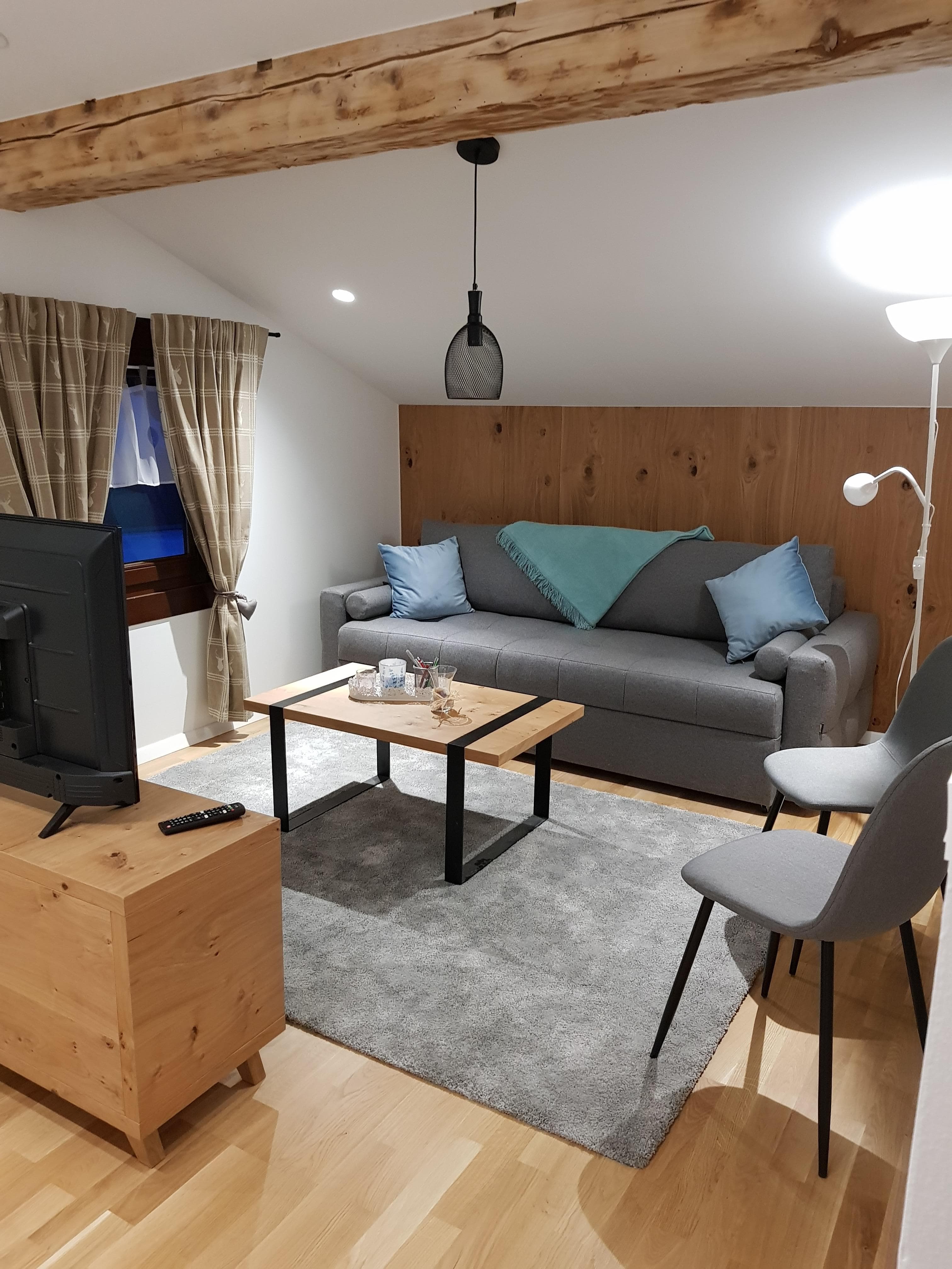 Apartments Kapelln · Panorama - A cozy bedroom with a large bed, featuring a grey upholstered headboard and decorative pillows. The room has wooden floors, white walls, and exposed wooden beams in the ceiling. Curtains cover a window, and a radiator is visible. Pillows on the bed have a 'Keep Calm and Drink Champagne' inscription.