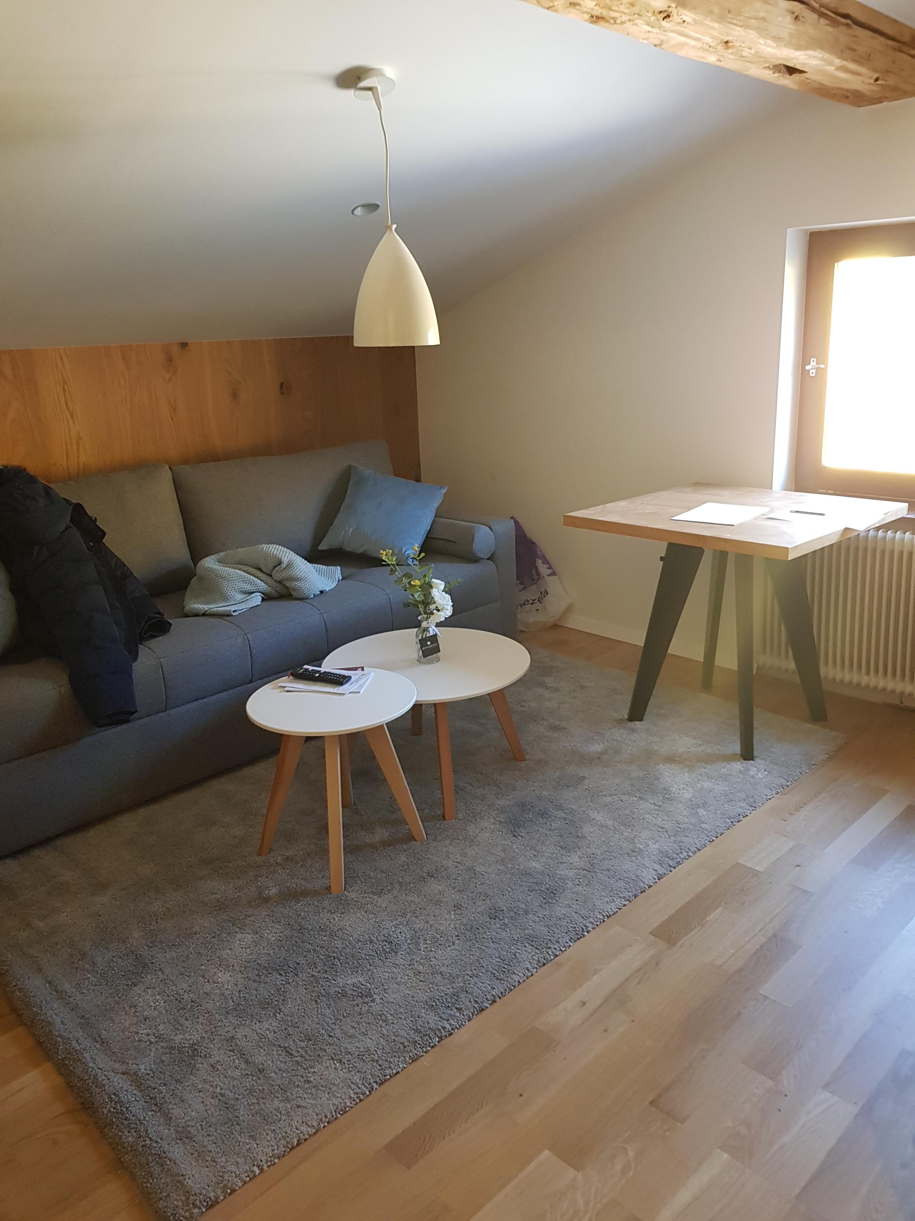 Apartments Kapelln · Buchensteinwand - A bedroom with a large wooden bed frame and a grey upholstered headboard. White bedding is neatly arranged. A bedside table with a lamp sits to the right. The walls are paneled with wood, and exposed wooden beams are visible on the ceiling.